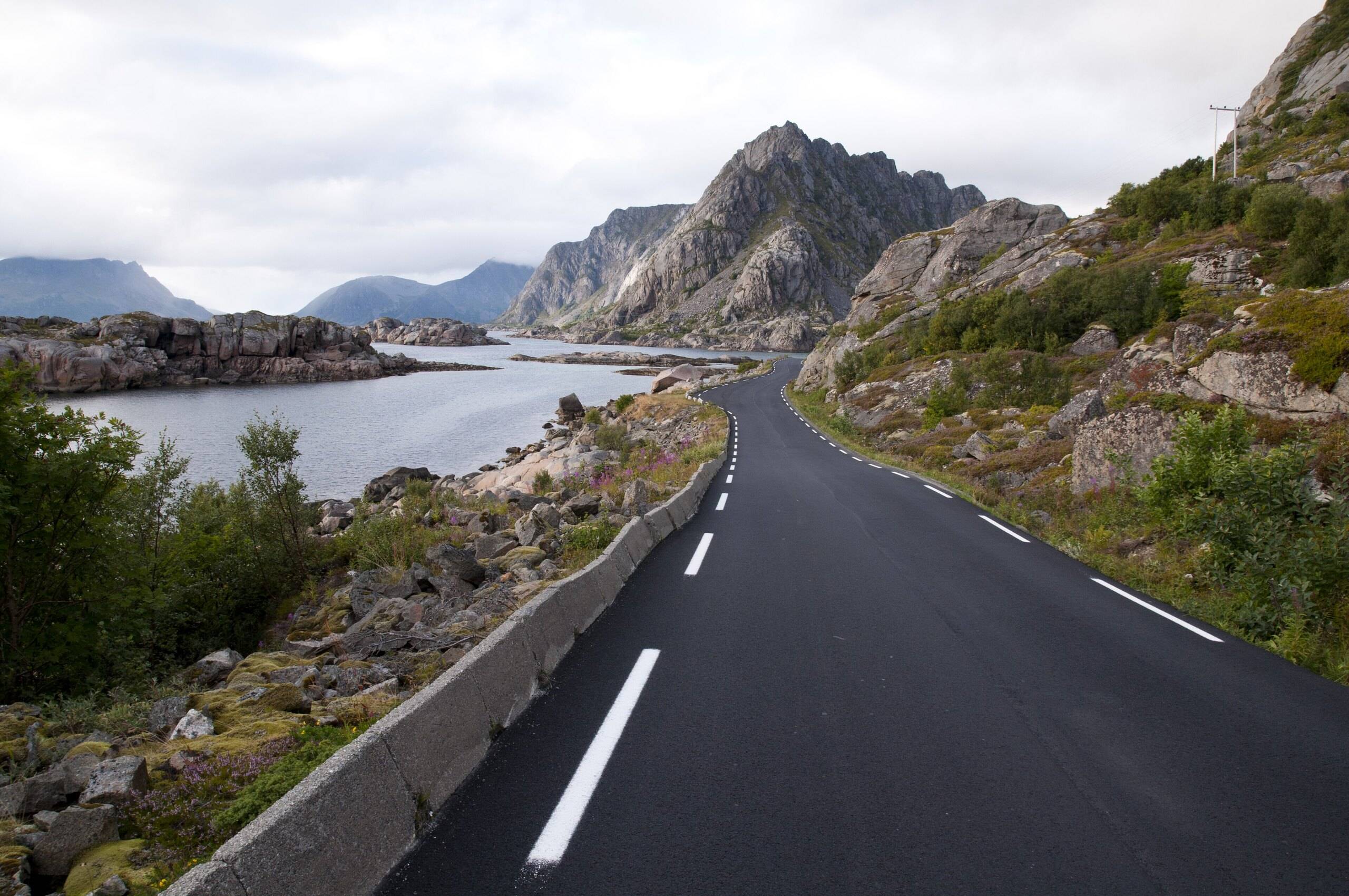 A two-lane road curving along the coast towards steep mountains in Lofoten